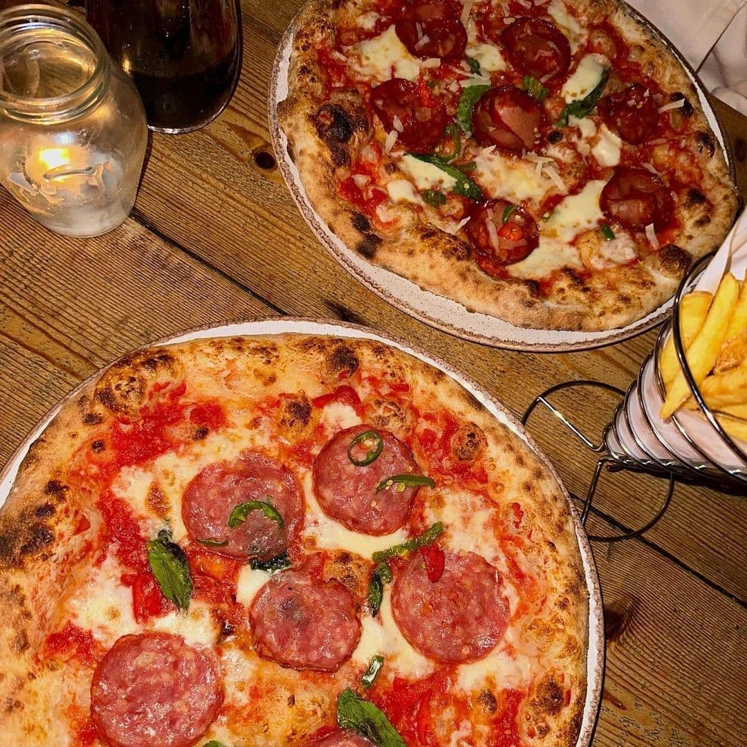 WOOD FIRE DINE Pizzeria Artisanal Pizzas Candlelight Dinner Freshly baked pizzas with toppings on wooden tables at WOOD FIRE | DINE Pizzeria, accompanied by a side of golden fries in a metal basket and a candlelit ambiance.
