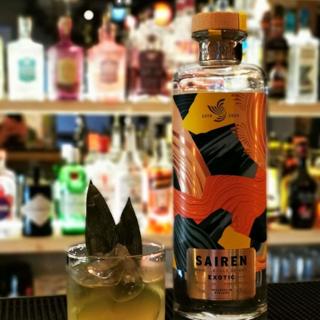 WOODFIRE DINE Pizzeria Exotic Bottle Cocktail Bar Atmosphere Close-up of a cocktail glass with ice and a clear beverage garnished with dark leaves, in front of a blurred array of colorful bottles at WOOD FIRE | DINE Pizzeria. A prominently displayed bottle labeled 'SAIREN Exotic' with a stylish design is in focus.