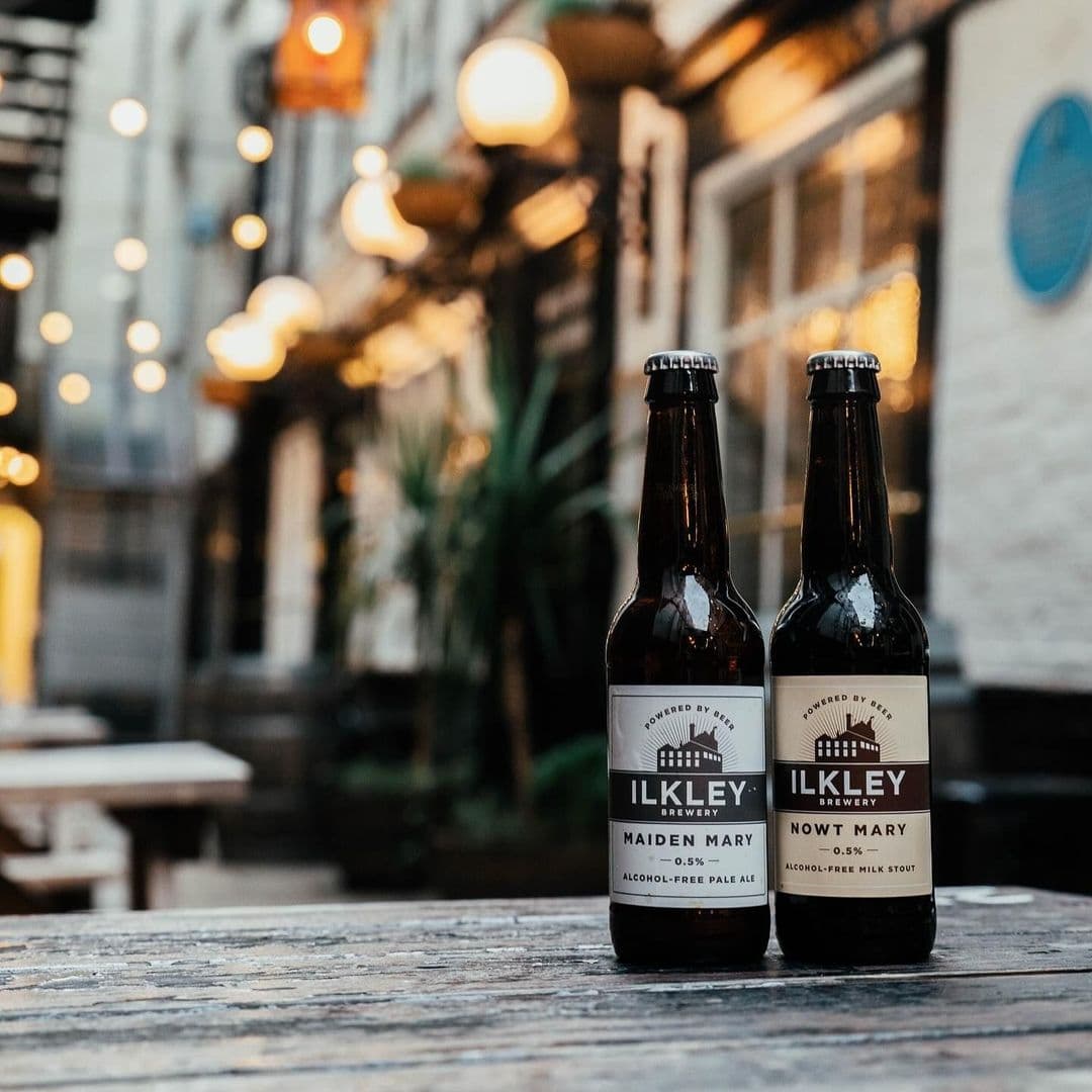 Whitelocks Ale House Craft Beer Bottles Evening Ambiance Two bottles of Ilkley Brewery beer on a wooden table at Whitelock's Ale House, with warm ambient lighting and blurred background.
