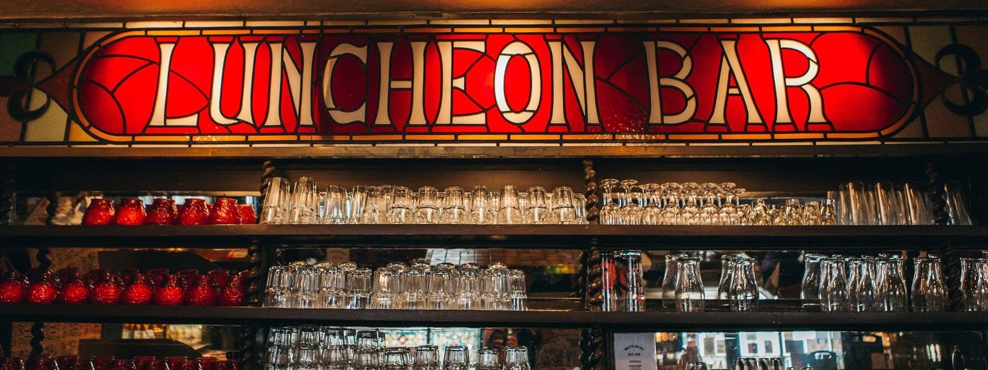 Interior of Whitelock's Ale House showcasing a stained glass sign reading "LUNCHEON BAR" above a shelf with neatly arranged glassware and red teacups.