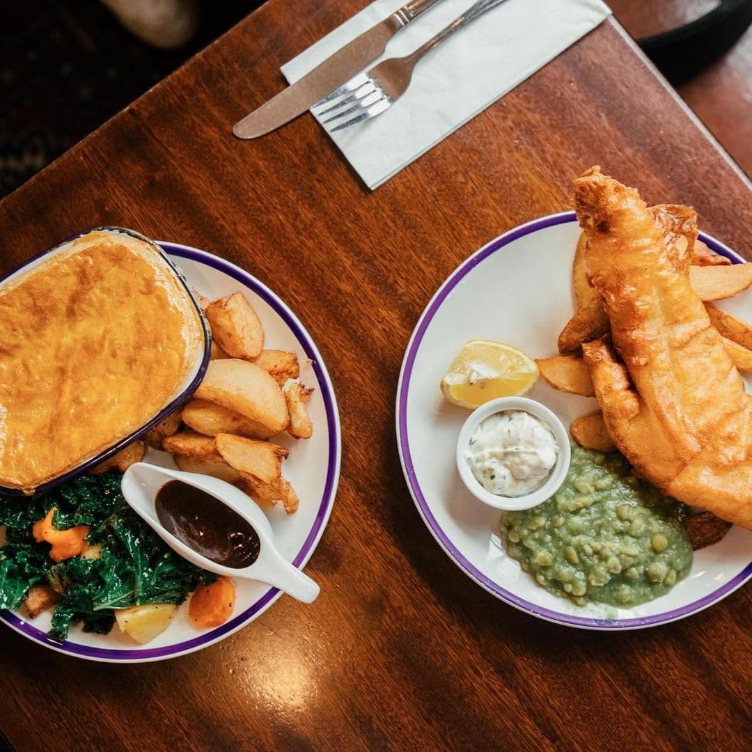 Whitelocks Ale House Traditional British Pub Food Traditional British cuisine served at Whitelock's Ale House, featuring a plate of fish and chips with a side of mushy peas and tartar sauce, and a pot pie with a golden crust, accompanied by roasted potatoes and steamed greens.