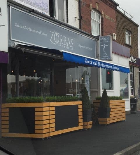 Zorbas_Bar_and_Grill_Street_View_Mediterranean_Cuisine Exterior view of Zorbas Bar and Grill, featuring a modern facade with signage, outdoor seating area with wooden planters, and large windows reflecting the street.