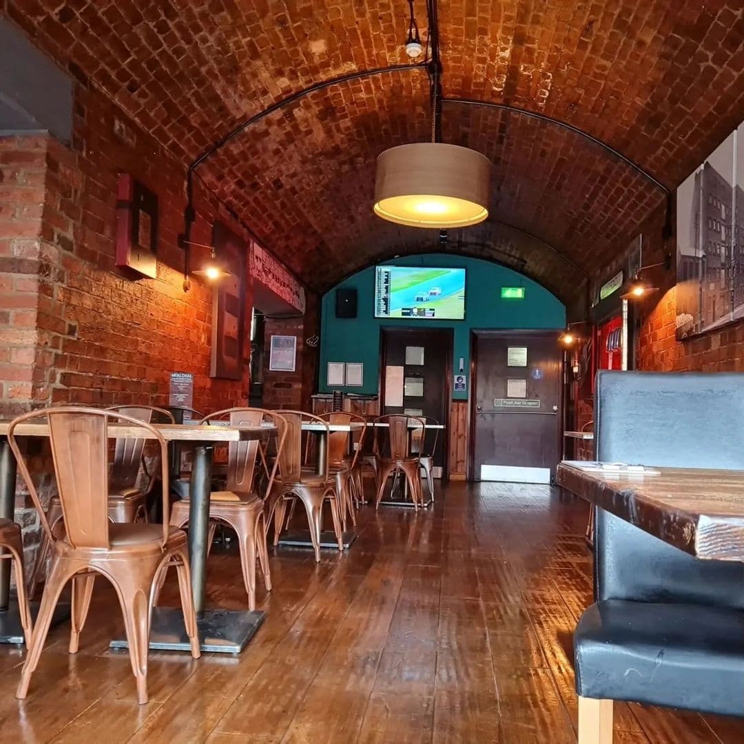 Aire_Bar_Vaulted_Ceiling_Brick_Interior_Empty_Tables Interior of Aire Bar featuring arched brick ceiling, wooden tables and chairs, and a television screen on the wall.