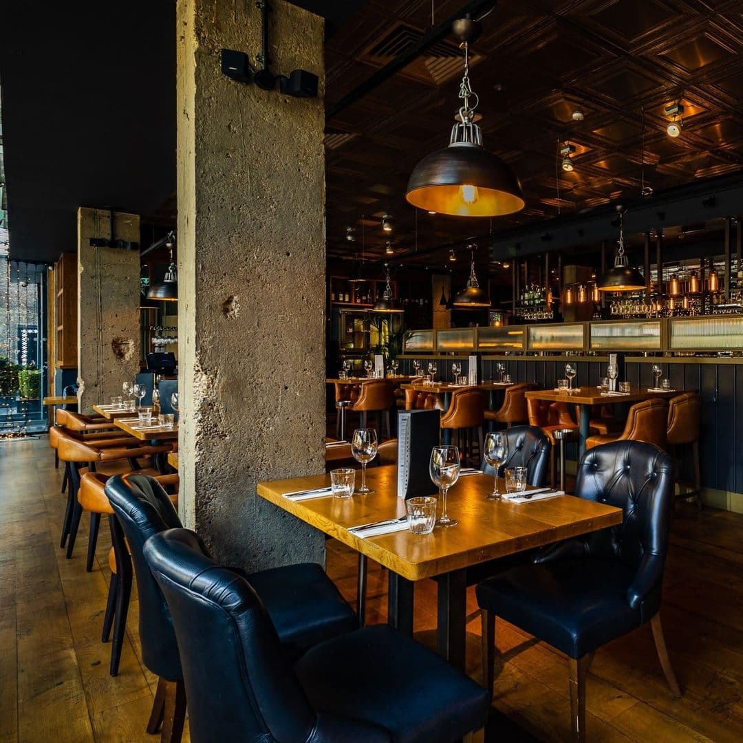 Blackhouse Elegant Dining Ambiance Wooden Tables Industrial Lights Interior of Blackhouse restaurant with wooden tables set for dining, black leather chairs, pendant lighting, and a bar with shelves of bottles in the background.