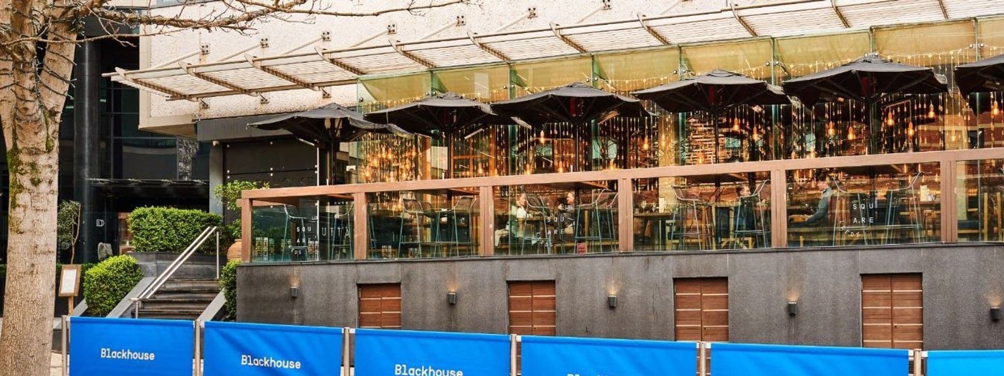 Exterior view of Blackhouse restaurant with glass facade and outdoor umbrellas, featuring warm interior lighting and modern architecture.