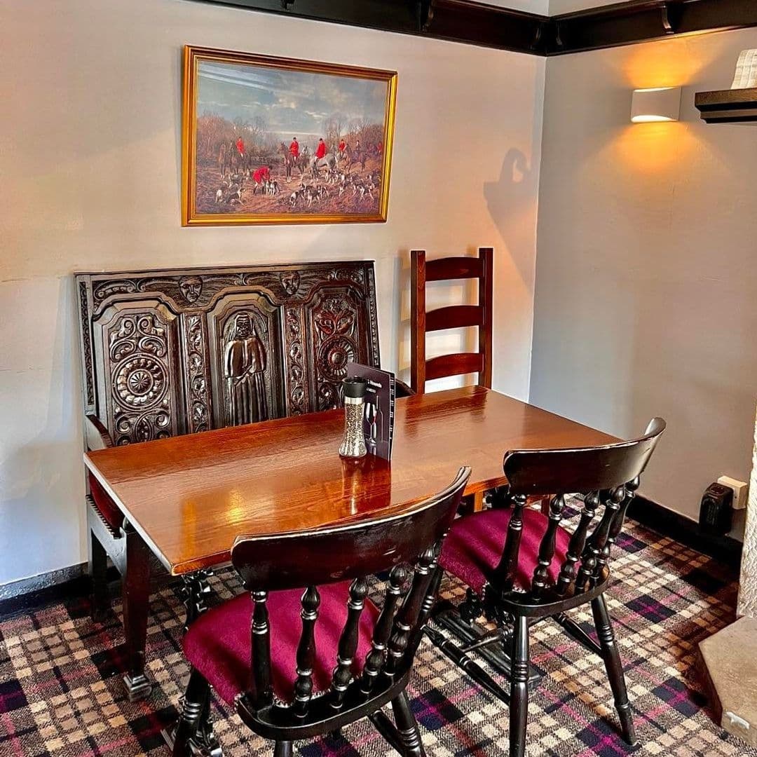 Fox and Hounds Cozy Traditional Interior Hunting Artwork Cozy dining area at Fox & Hounds with traditional wooden furniture, plush burgundy seats, a classic fox hunt painting on the wall, and ornate carved wood bench.