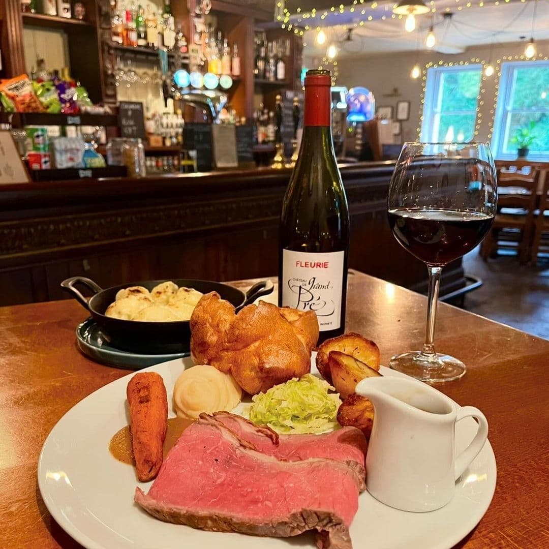 Ilkley Moor Vaults Traditional British Roast Dinner Wine Traditional British roast dinner with slices of roast beef, Yorkshire pudding, roasted potatoes, and vegetables, served with gravy on a white plate, accompanied by a bottle of Fleurie red wine and a glass, at Ilkley Moor Vaults. The background features a cozy pub setting with a wooden bar and festive lights.