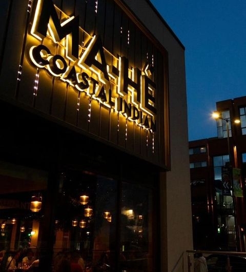 Ma Hé Coastal Indian Restaurant Evening Ambience Exterior view of Ma-Hé Coastal Indian restaurant at dusk with illuminated sign and string lights, patrons dining inside visible through the windows.