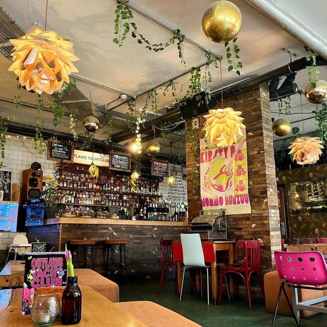 Outlaws_Yacht_Club Hip_Bar_Interior_Decor Vintage_Style Interior of Outlaws Yacht Club with eclectic decor, featuring hanging paper lanterns, trailing green plants, and disco balls. A well-stocked bar with a variety of bottles is visible in the background, and colorful chairs are scattered around wooden tables in the foreground.