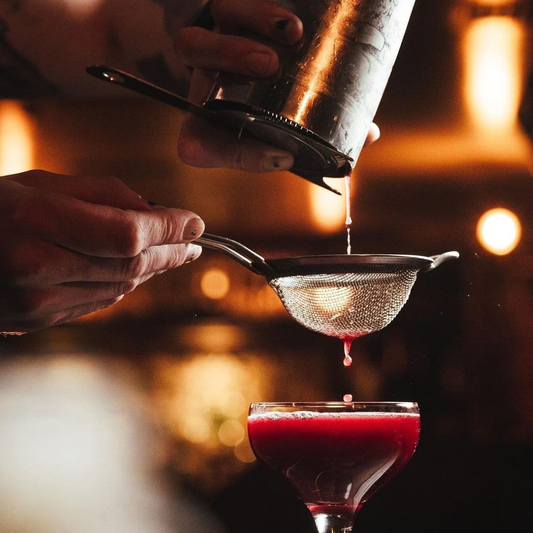 Smokestack_Cocktail_Pouring_Fine_Strainer_Ambient_Lighting Bartender pouring a red cocktail through a strainer into a glass at Smokestack, with a warm, dimly lit ambiance in the background.