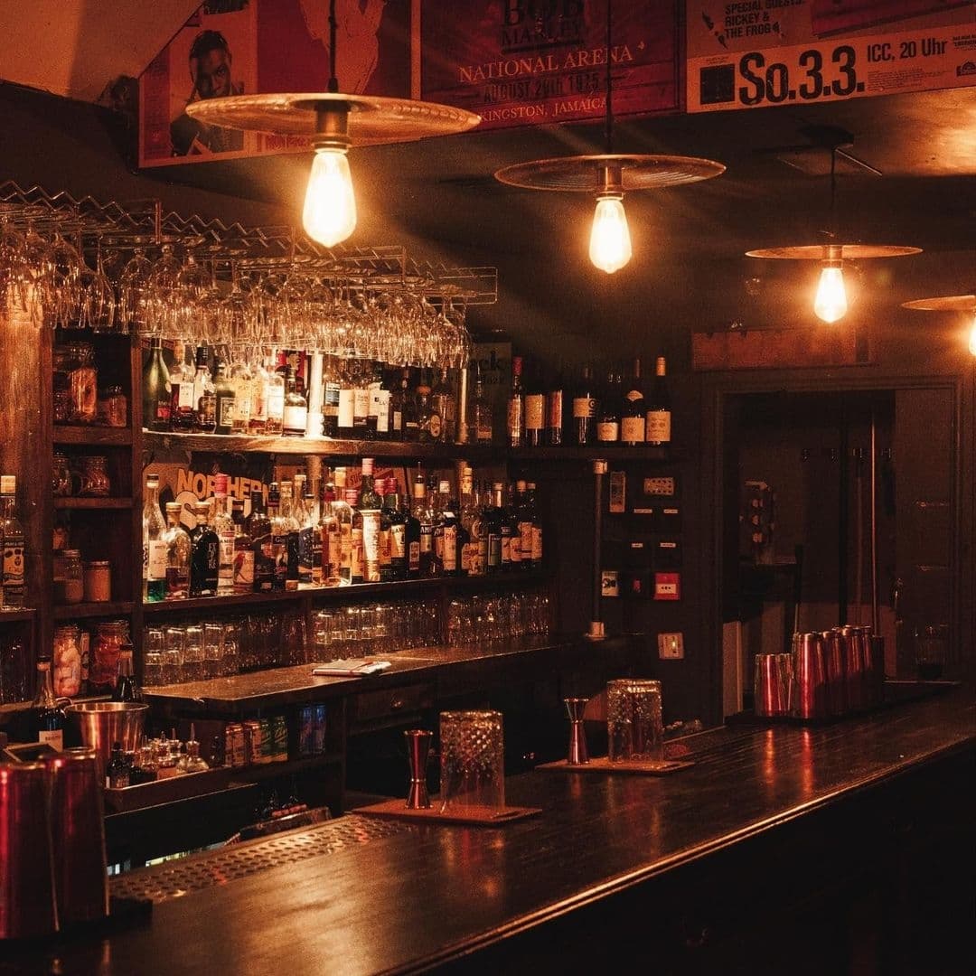 Smokestack_VintageBarAmbiance_WarmLighting_WoodenInterior Interior of Smokestack bar with dim lighting, showcasing an array of bottles on wooden shelves, hanging wine glasses, and vintage pendant lights.
