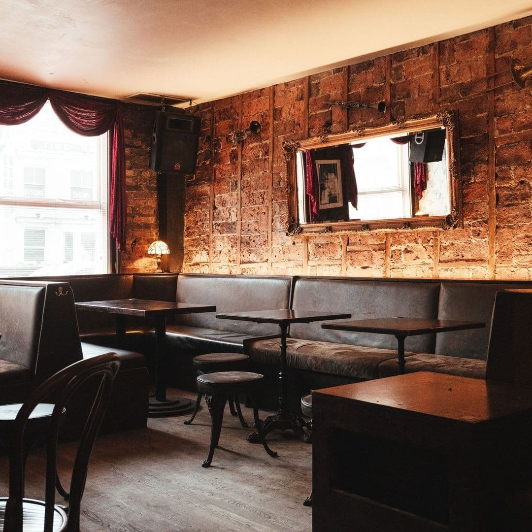 Smokestack_VintagePubInterior_RusticBrickWalls_ComfySeating Interior of Smokestack featuring rustic brick walls, cozy seating with leather benches and wooden chairs, ambient lighting, and a large mirror reflecting the window.