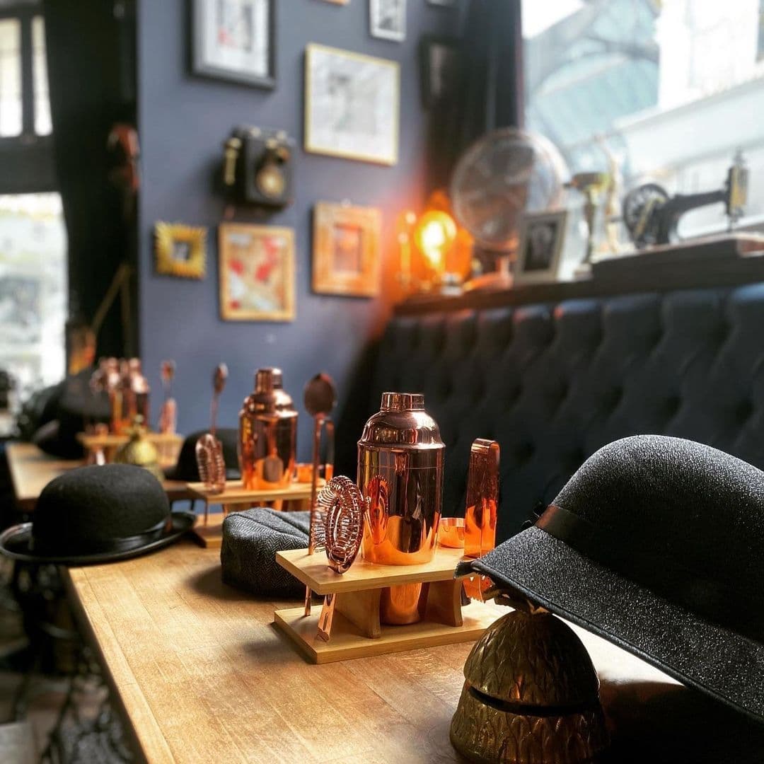 TailorsGin_CozyBarAmbiance_CopperCocktailTools_VintageDecor Interior of Tailors Gin with elegant copper cocktail shakers on a wooden bar, vintage framed pictures on dark blue walls, and stylish black bowler hats adding to the ambiance.