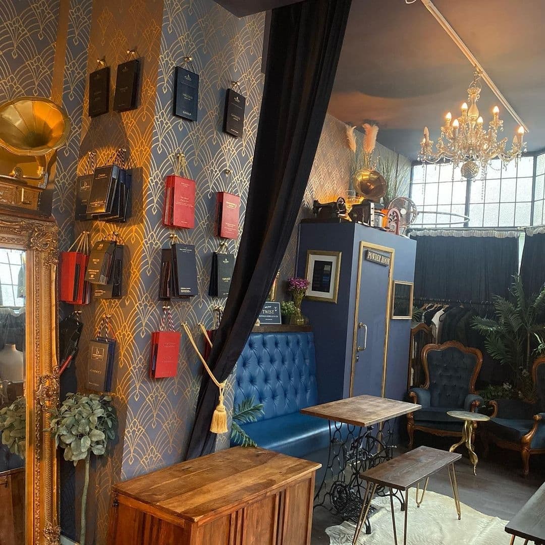 TailorsGin_VintageChicInterior_ElegantDecor_BookshelfWall Elegant interior of Tailors Gin with a vintage vibe featuring blue tufted seating, ornate gold-framed mirror, and book-themed wall decor under a classic chandelier.