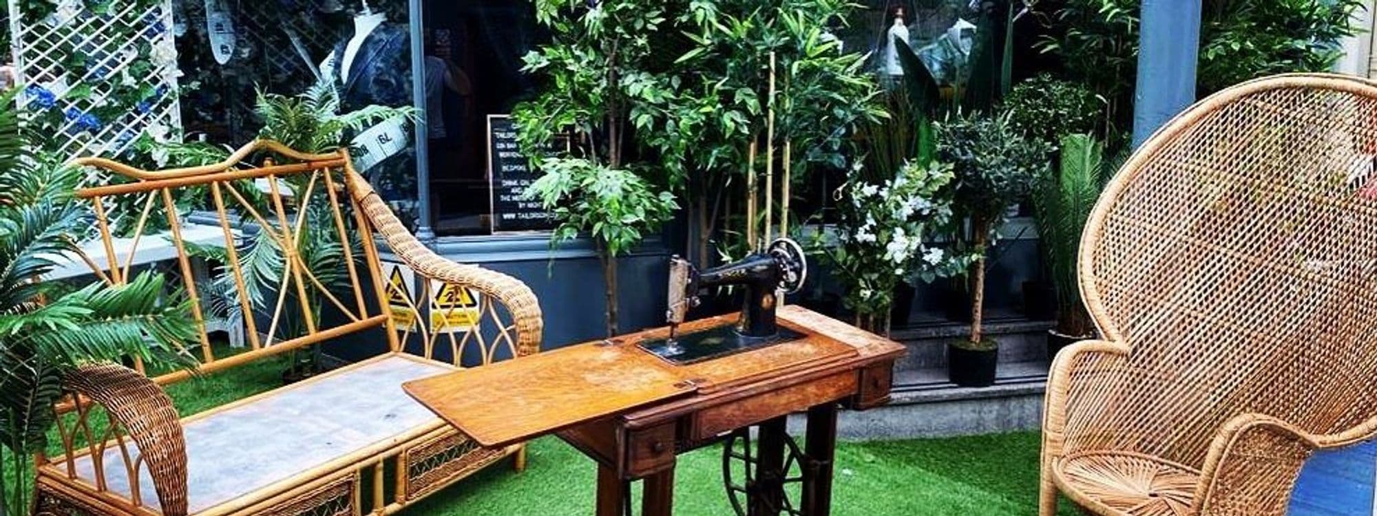 Outdoor seating area at Tailors Gin with wicker chairs, a vintage sewing machine table, and lush green plants.