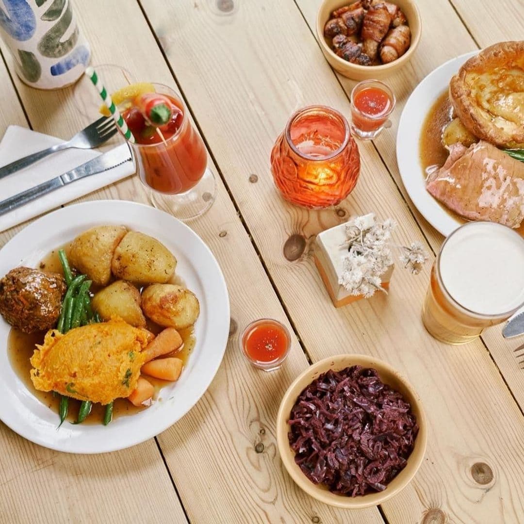 The Brunswick Traditional English Breakfast Hearty Meal Hearty meal spread on a wooden table at The Brunswick, featuring roasted potatoes, green beans, carrots, a meatball, red cabbage, sausages, French toast with syrup, a glass of beer, and a Bloody Mary cocktail.