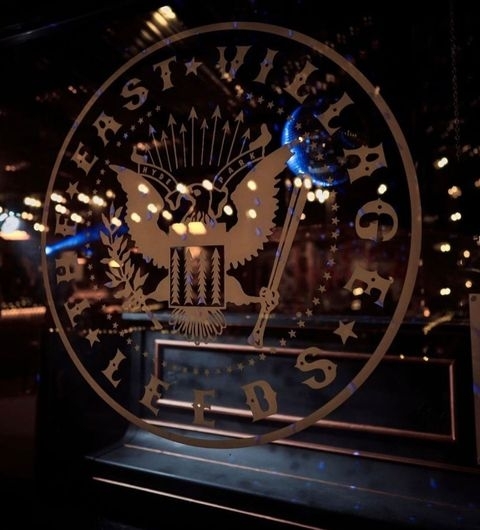 The East Village Emblem Nightlife Bokeh Lights Decorative window decal featuring an eagle and stars with the text "The East Village" at a local establishment, illuminated by warm ambient lighting.