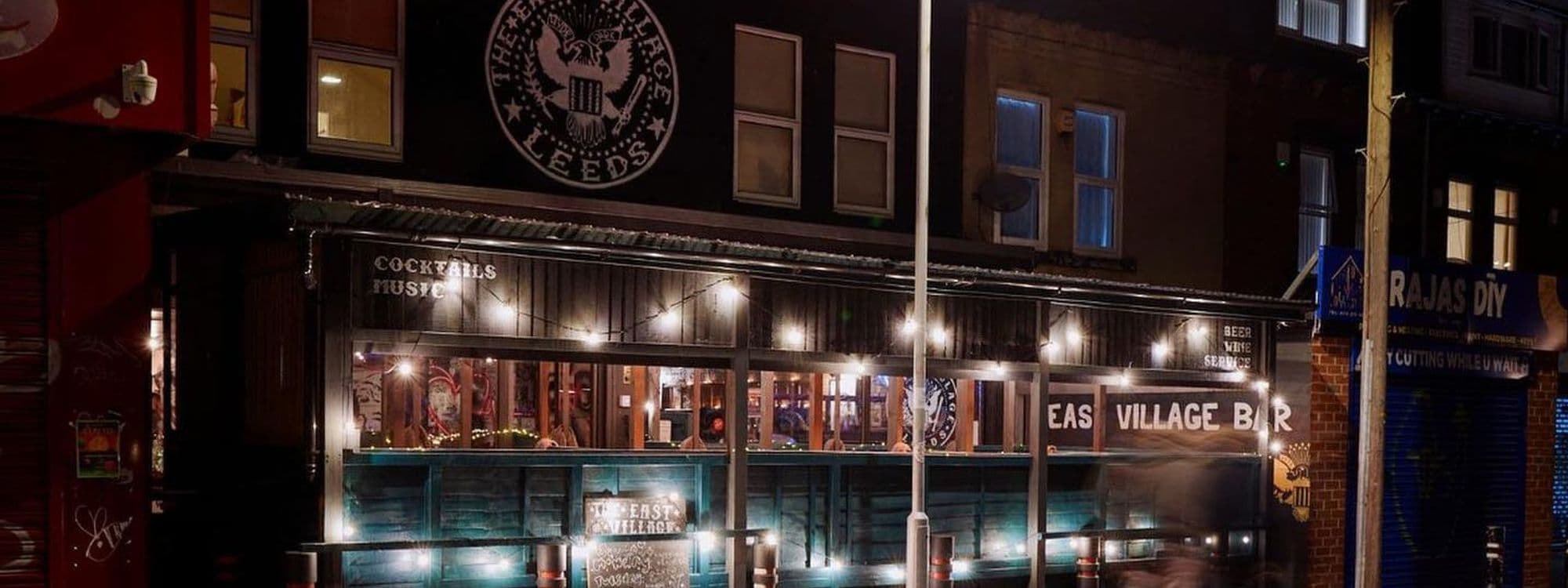 Nighttime view of The East Village Bar with illuminated string lights and a prominent eagle emblem above the entrance, reflecting the vibrant nightlife and social scene.