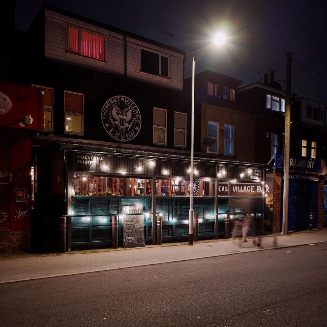The East Village Nightlife Bar Exterior Street View Nighttime view of The East Village bar with illuminated windows and a person walking by on the sidewalk.