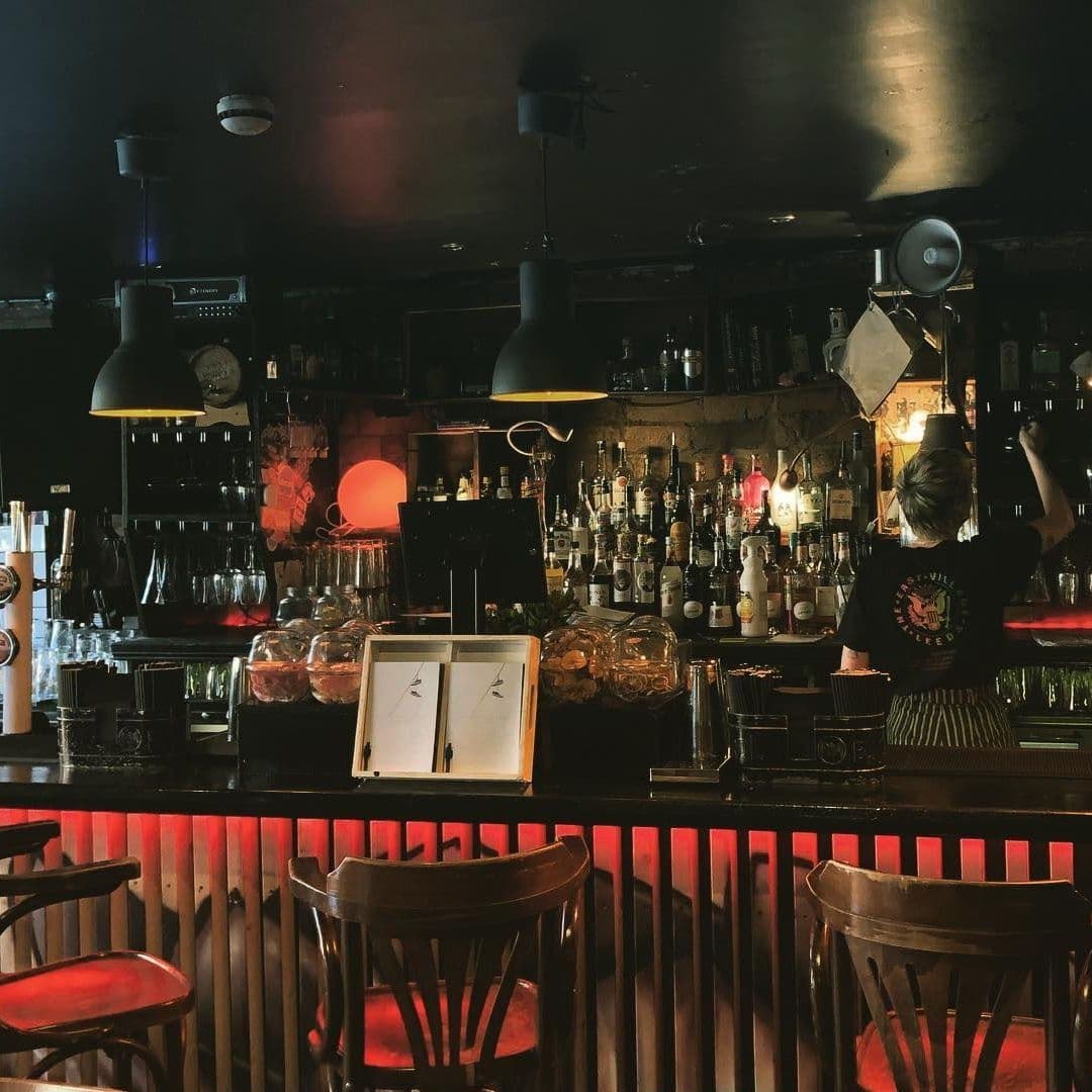 The East Village cozy bar interior mood lighting Interior of a cozy bar in The East Village with dim lighting, featuring a bartender at work, an assortment of bottles on shelves, pendant lamps, and wooden bar stools.