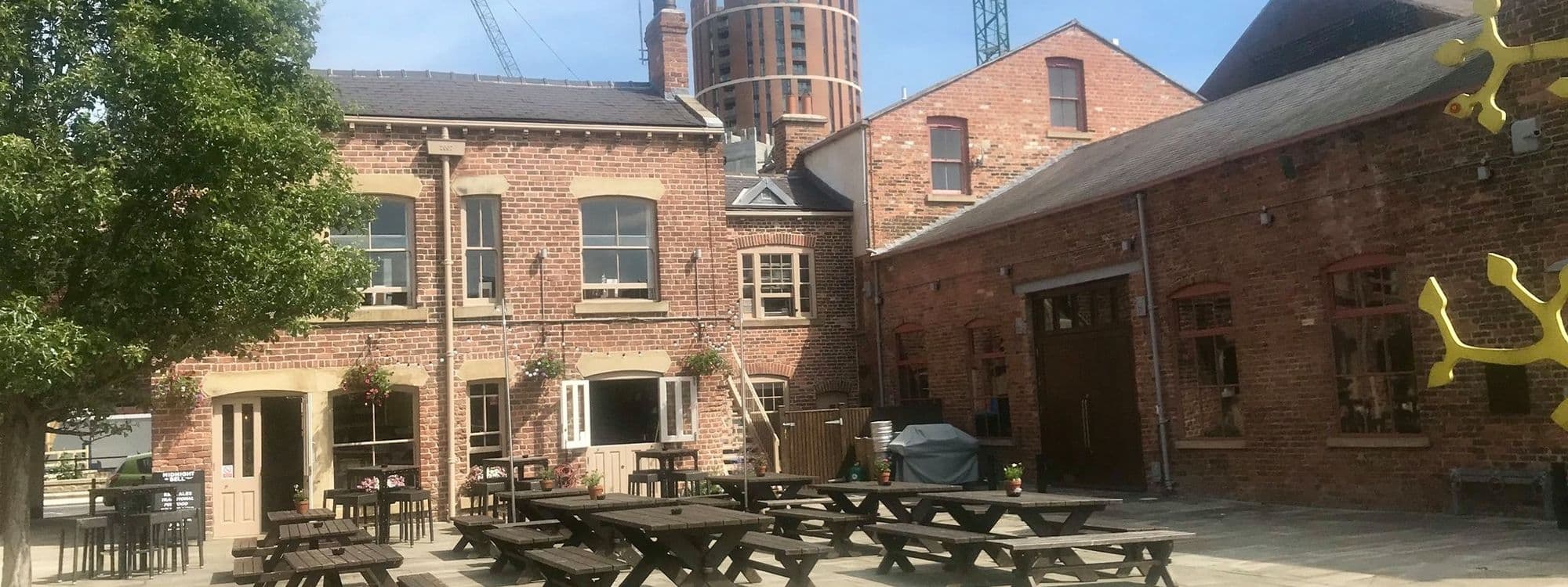 Outdoor seating area at The Midnight Bell with wooden picnic tables, brick buildings, and clear blue sky.