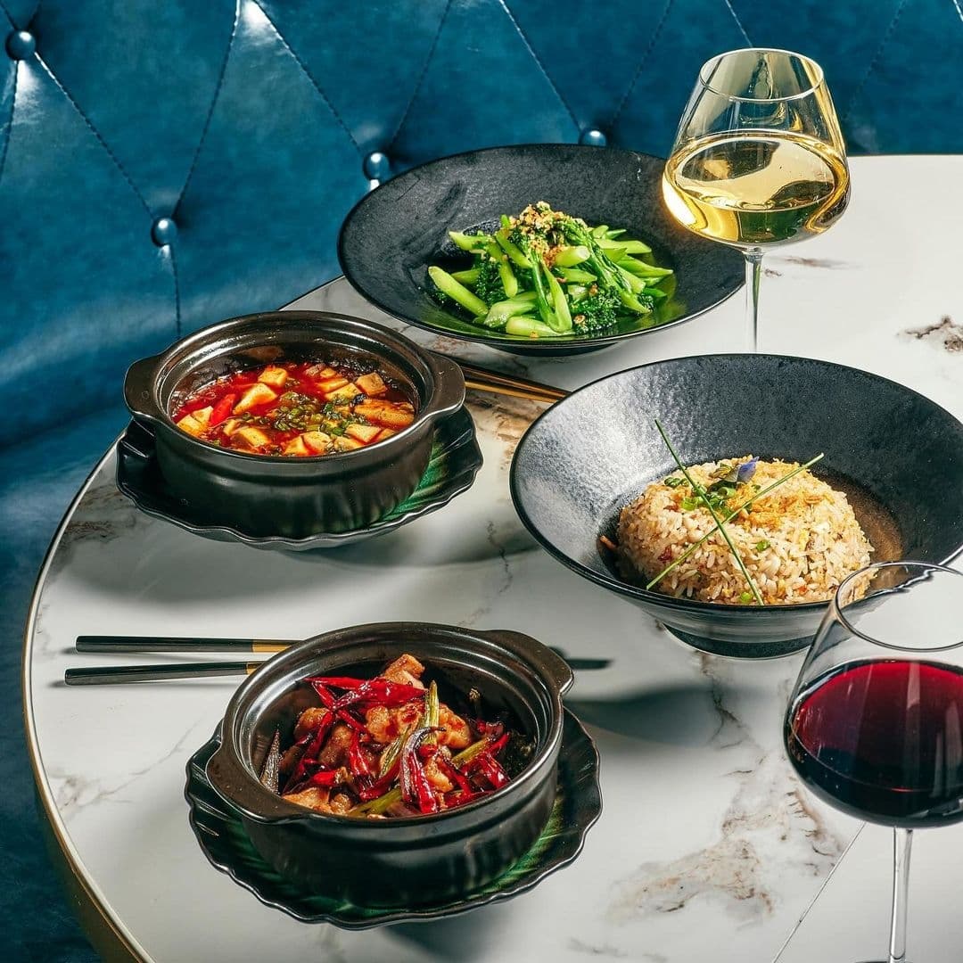 Blue Pavilion_Gourmet Dining Experience_Elegant Setting Elegant dining setup at Blue Pavilion featuring a variety of Asian cuisine dishes and a glass of white wine and red wine on a marble table with a blue tufted booth in the background.