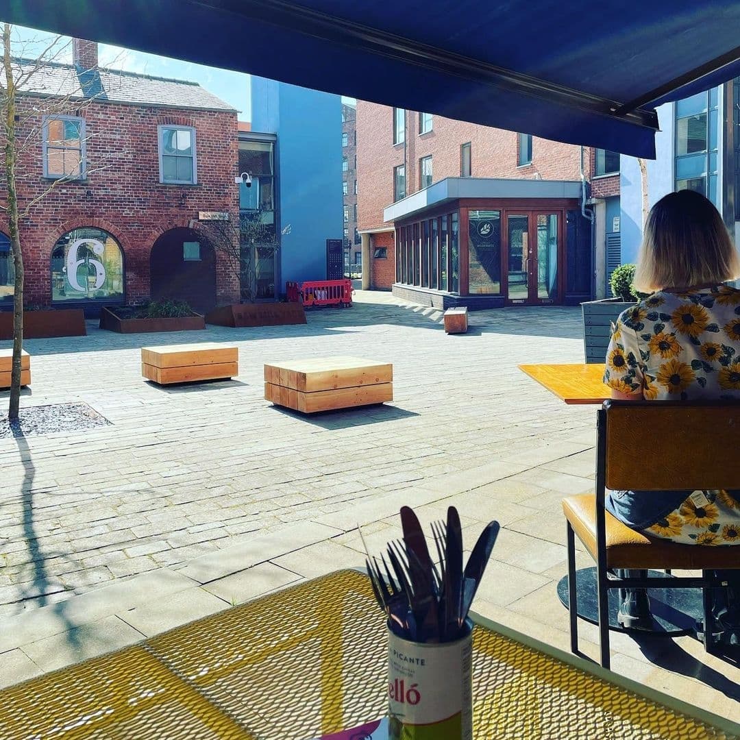 Bomba Paella Outdoor Dining Sunny Day Relaxed Atmosphere Outdoor seating at Bomba Paella with a view of a sunny courtyard, wooden benches, a woman in a floral dress, and brick buildings.