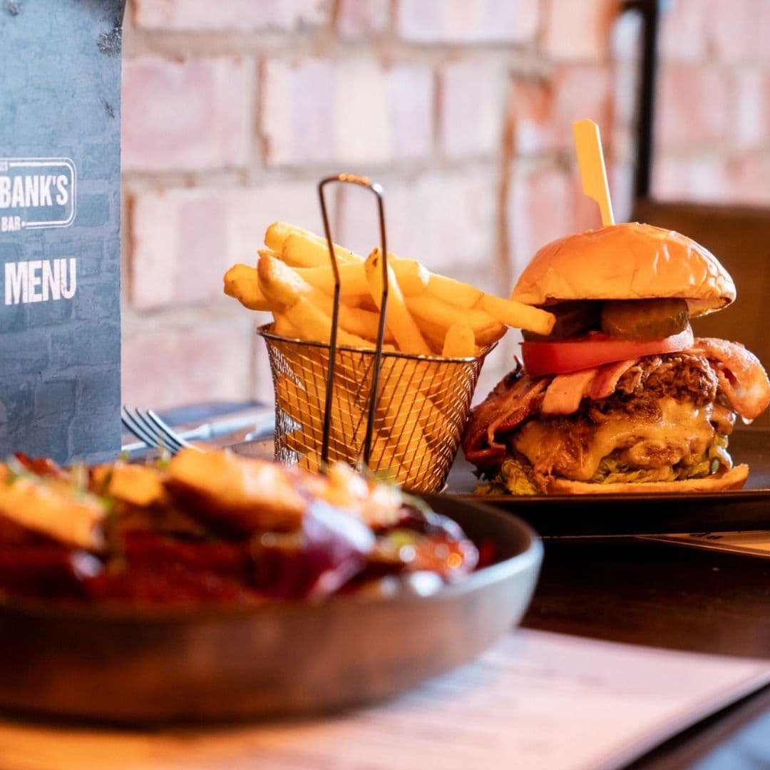 Bricklebanks Bar Kitchen Gourmet Burger Fries Appetizing Plate Savory meal at Bricklebank's Bar & Kitchen featuring a stacked burger with cheese, tomato, and crispy onions, accompanied by a basket of golden fries, with a menu visible in the background.