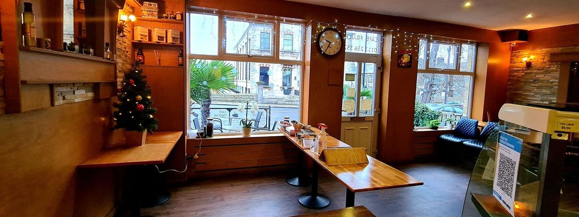 Interior of Cafe Fraiche with wooden tables and benches, a Christmas tree, and a view of the street through large windows. Decorative lights and a stone accent wall create a cozy atmosphere.