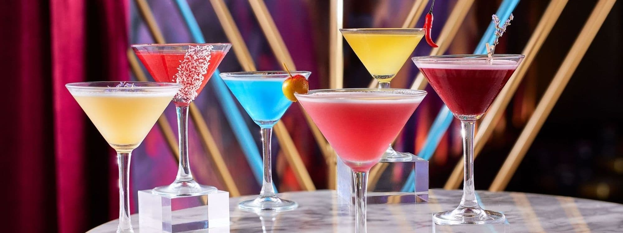 Assorted colorful cocktails on a marble bar counter at Dirty Martini, with a vibrant backdrop of red curtains and abstract art.
