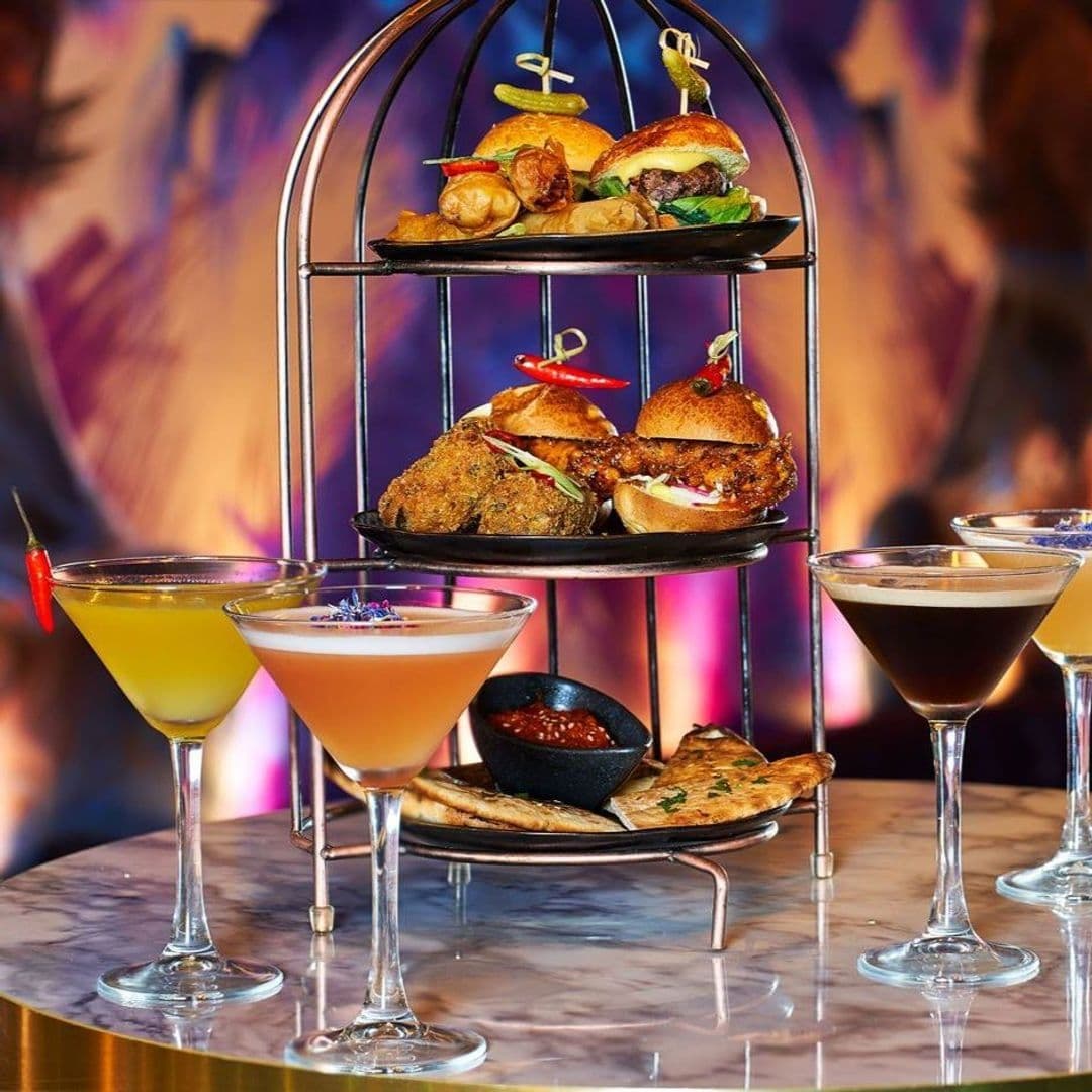 DirtyMartini_Cocktails_FoodPlatter_ElegantPresentation Elegant food and cocktail presentation at Dirty Martini, featuring a tiered stand with gourmet sliders, stuffed jalapeños, and other appetizers, accompanied by an assortment of colorful martinis on a marble table with a vibrant, blurred background.