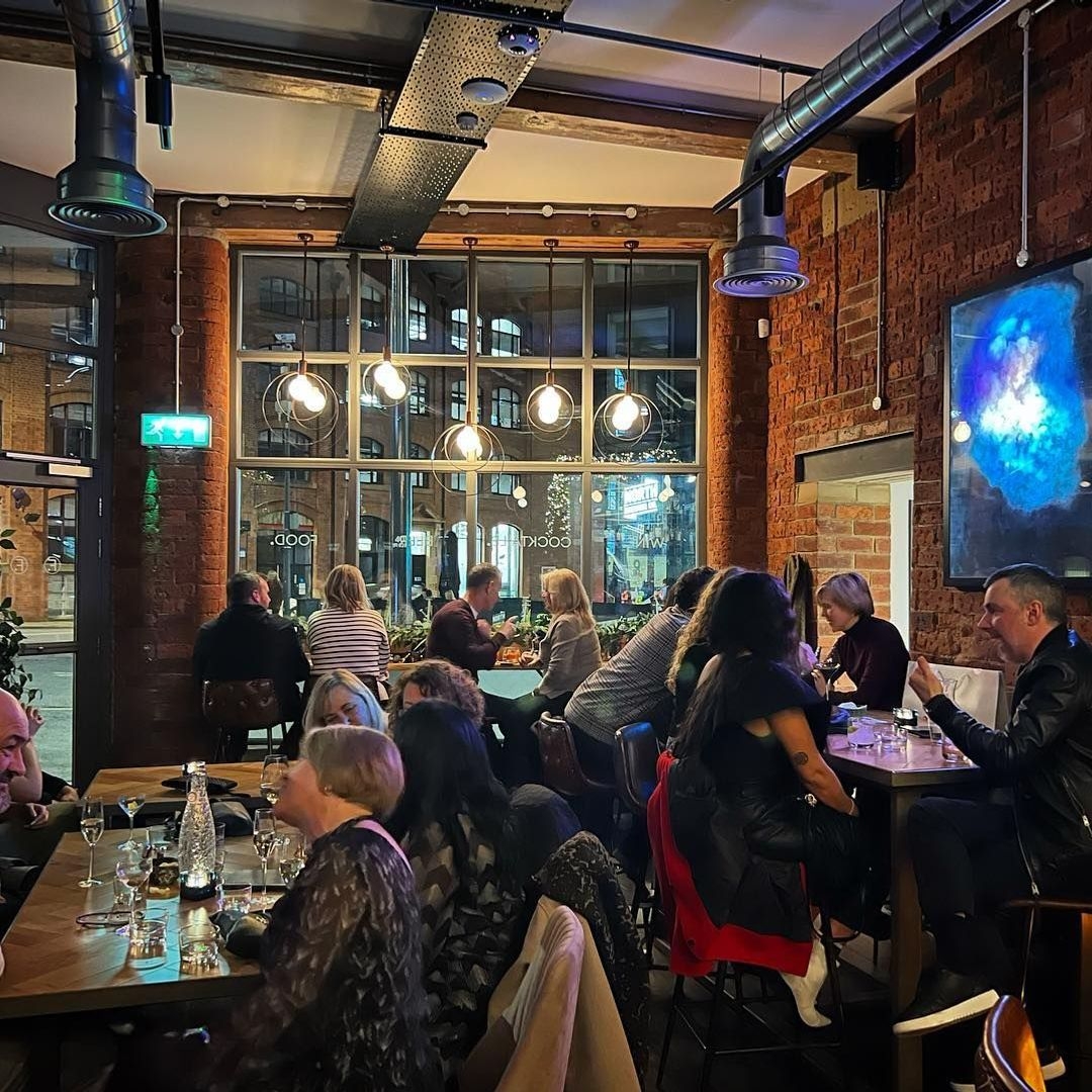 Farrands_Bar_Cozy_Ambiance_Brick_Walls_Guests_Dining Patrons enjoying a lively atmosphere inside Farrands Bar, with exposed brick walls, modern hanging lights, and large windows offering a street view.