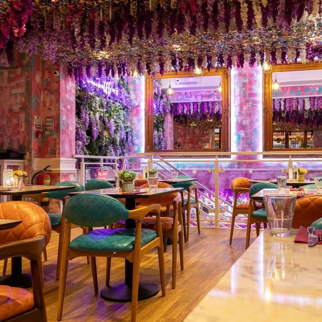 Fleur Restaurant and Bar Colorful Interior Floral Decor Vibrant Lighting Interior of Fleur Restaurant and Bar featuring vibrant floral decor with hanging purple flowers, eclectic furniture with teal and orange chairs, and illuminated pink neon lights.
