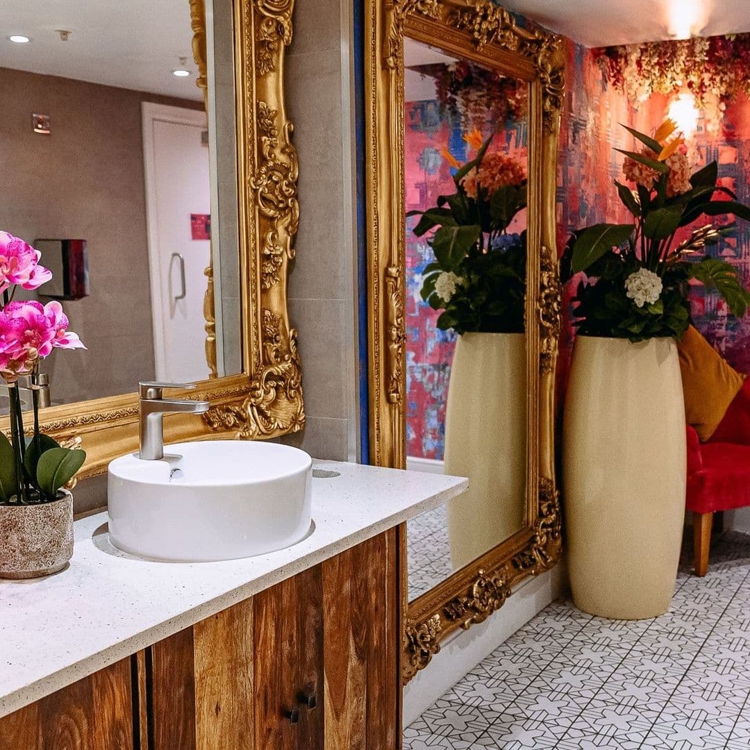 Fleur Restaurant and Bar Elegant Restroom Decor Gold Mirror Elegant bathroom interior at Fleur Restaurant and Bar with ornate gold-framed mirror, white basin on wooden vanity, and vibrant floral wallpaper.