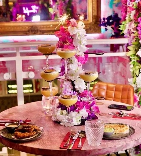 Fleur Restaurant and Bar_Elegant Cocktail Tower_Floral Decor_Dining Experience Alt text: "Elegant dining setup at Fleur Restaurant and Bar featuring a tower of champagne glasses adorned with flowers, surrounded by vibrant floral decor, with dishes and cutlery on a table ready for a luxurious meal."