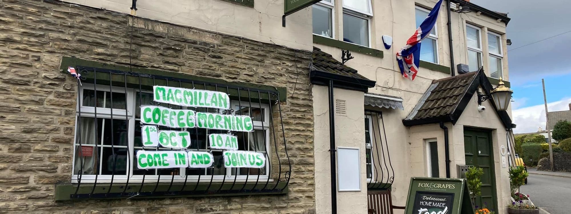 Exterior view of Fox & Grapes, featuring a stone facade with windows displaying a green banner for a Macmillan Coffee Morning event, a British flag, and a signboard advertising homemade food.