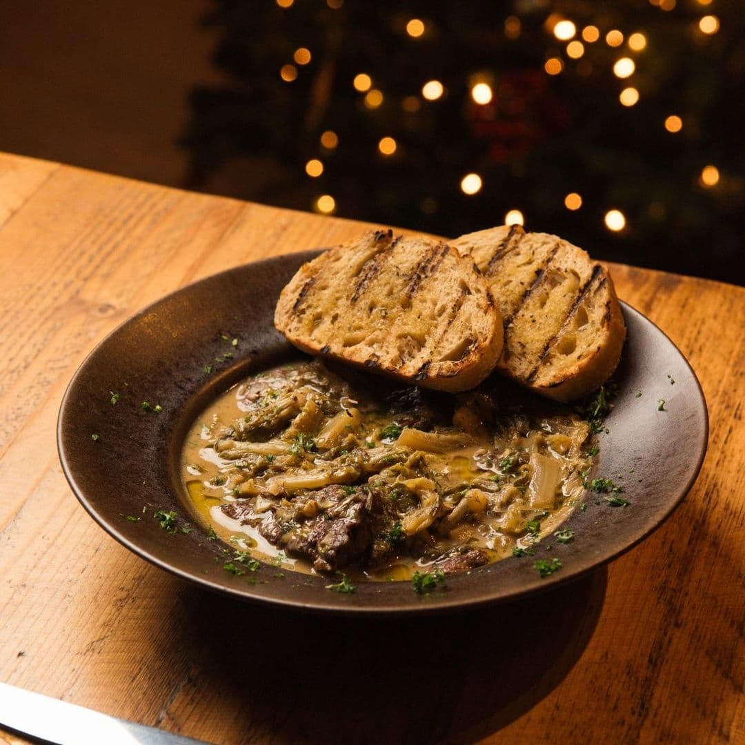 MYTHOS Chapel Allerton Cozy Winter Stew Toasted Bread Alt text: "Savory stew served at MYTHOS Chapel Allerton, with tender pieces of meat and vegetables, accompanied by two slices of grilled bread, presented on a dark ceramic plate against a backdrop of warm, twinkling lights."