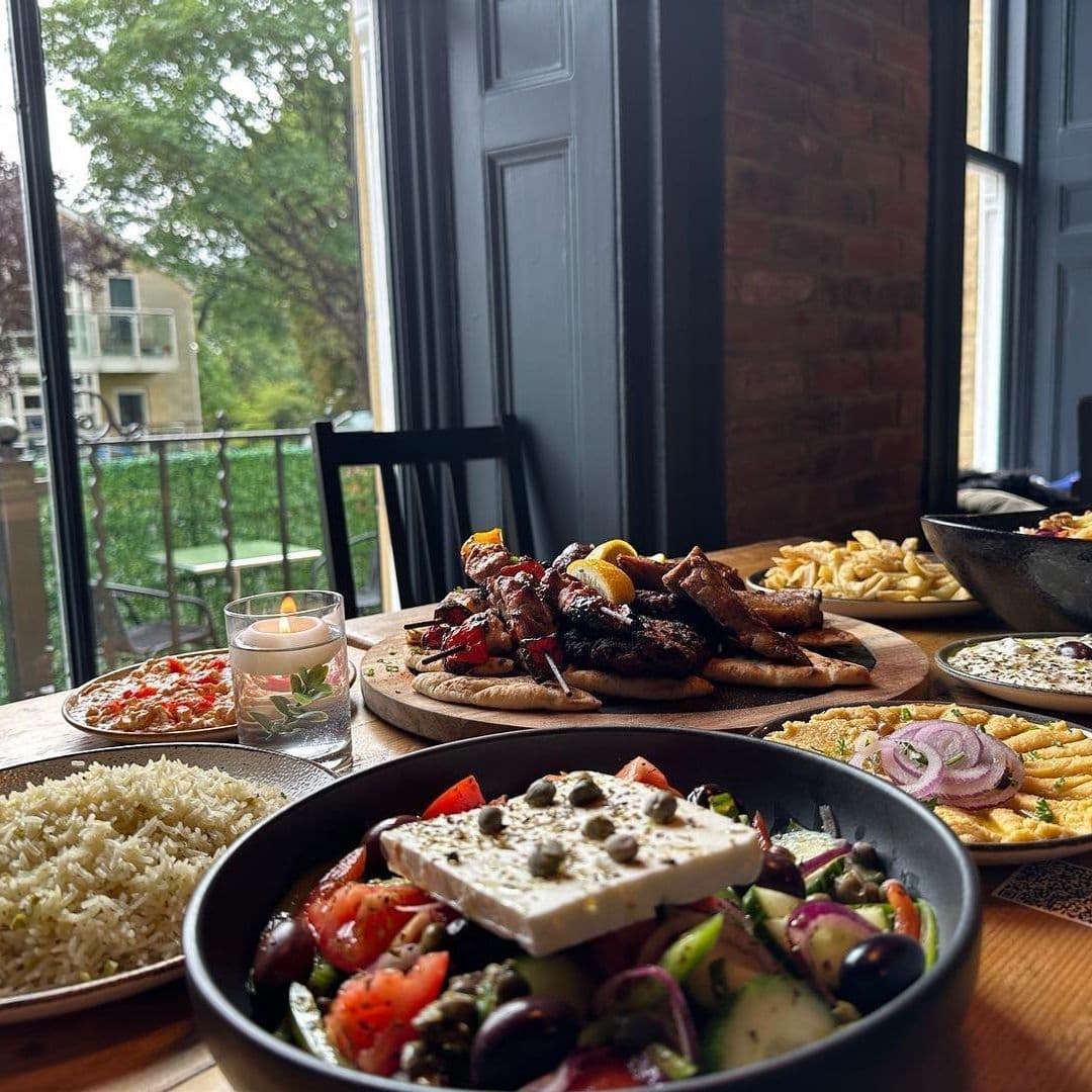 MYTHOS Chapel Allerton Greek Cuisine Dining Experience Assorted Greek dishes on a table at MYTHOS Chapel Allerton, featuring skewered meats, salad with feta, rice, and pasta, with a candlelit ambiance and a view of greenery outside the window.