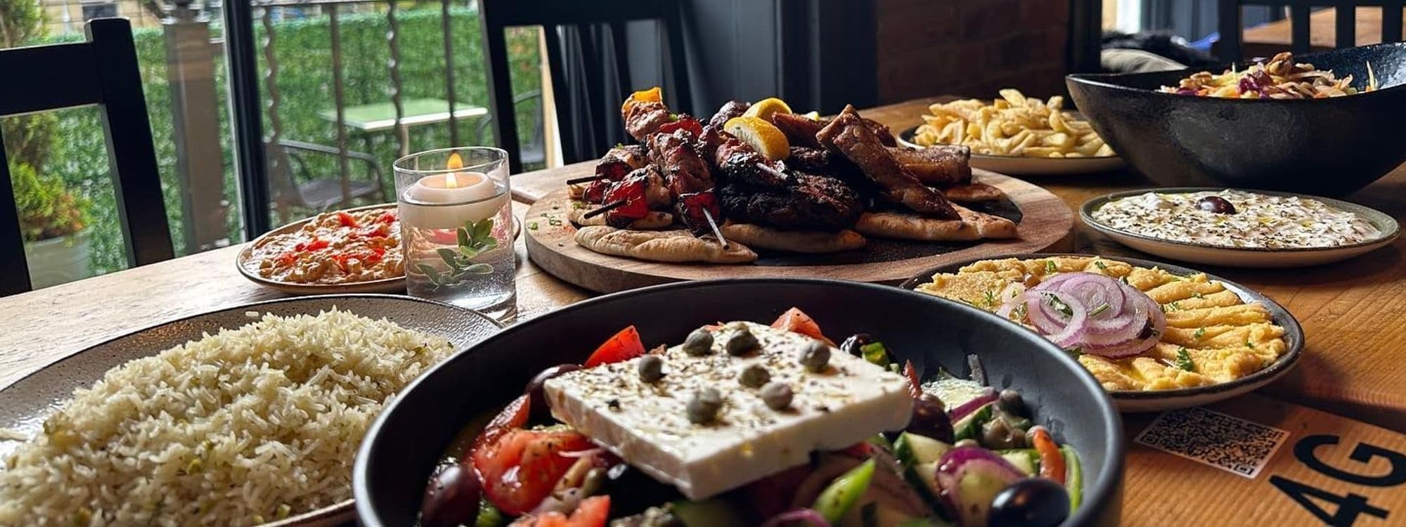 Assorted Greek dishes on a wooden table at MYTHOS Chapel Allerton, featuring skewered grilled meats, rice, Greek salad with feta cheese, pita bread, and dips, with a lit candle adding ambiance.