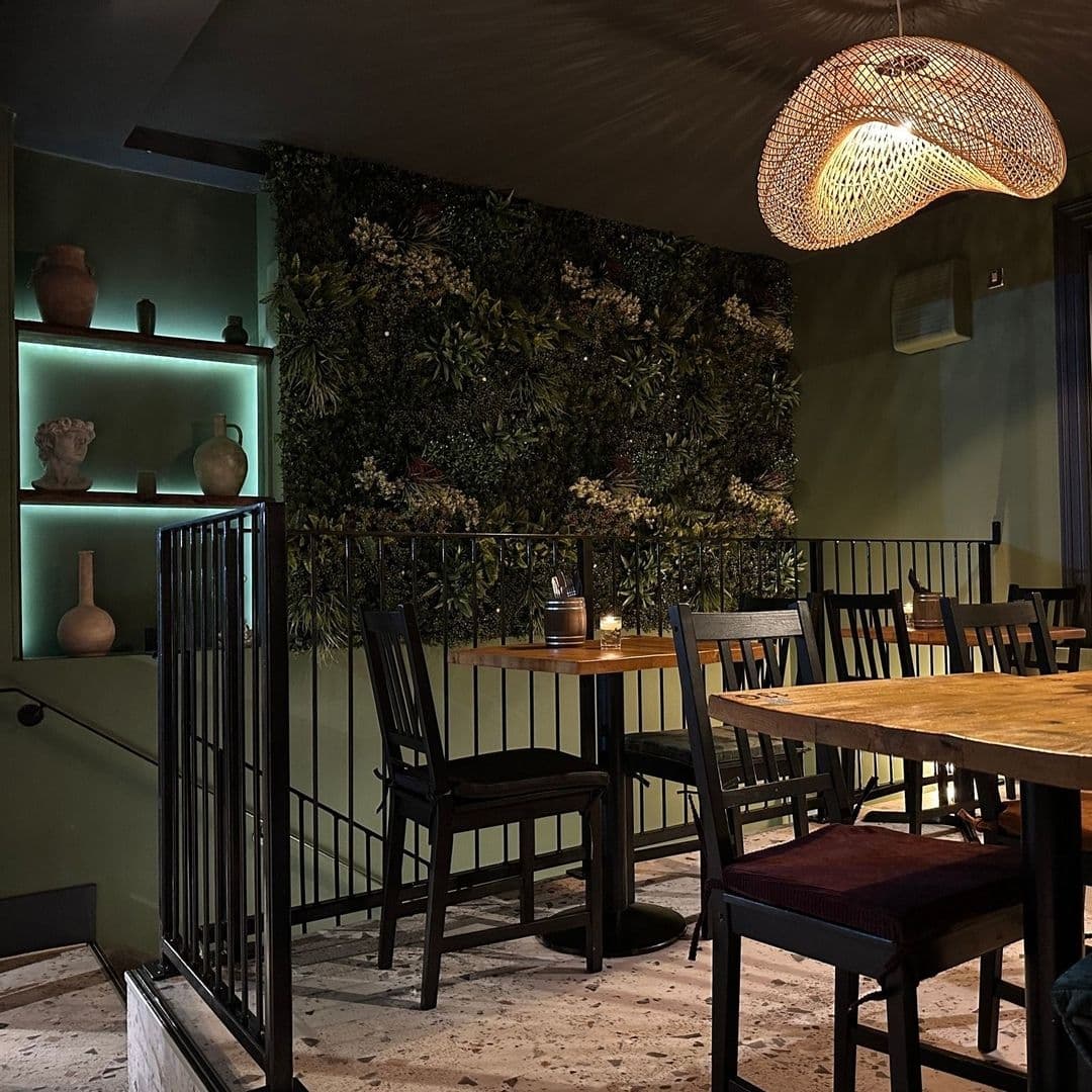 MYTHOS Chapel Allerton Intimate Dining Ambiance Greenery Interior of MYTHOS Chapel Allerton featuring a cozy dining area with wooden tables and chairs, ambient lighting from a woven pendant lamp, and a decorative green wall with plants.