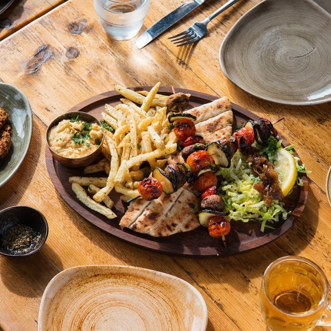 MYTHOS Chapel Allerton Mediterranean Cuisine Dining Experience Appetizing Greek meal served at MYTHOS Chapel Allerton, featuring skewered grilled vegetables and meat, seasoned fries, pita bread, and a side of hummus, with a glass of beer on a wooden table.