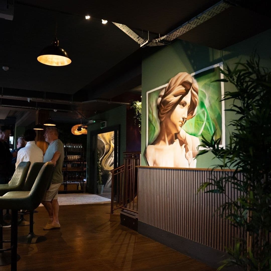 MYTHOS_Chapel_Allerton_Artistic_Interior_Decor_Patrons Interior of MYTHOS Chapel Allerton featuring patrons conversing, elegant green seating, and a large mural of a classical figure.