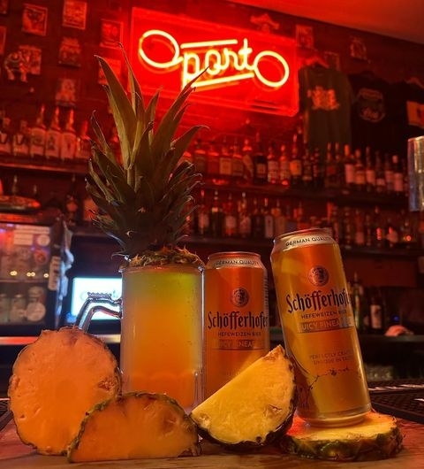 Oporto_NeonSign_PineappleBeer_BarAmbiance Alt text: "Chilled atmosphere in Oporto with a neon sign, fresh pineapple, and Schofferhofer wheat beer cans on a bar counter."