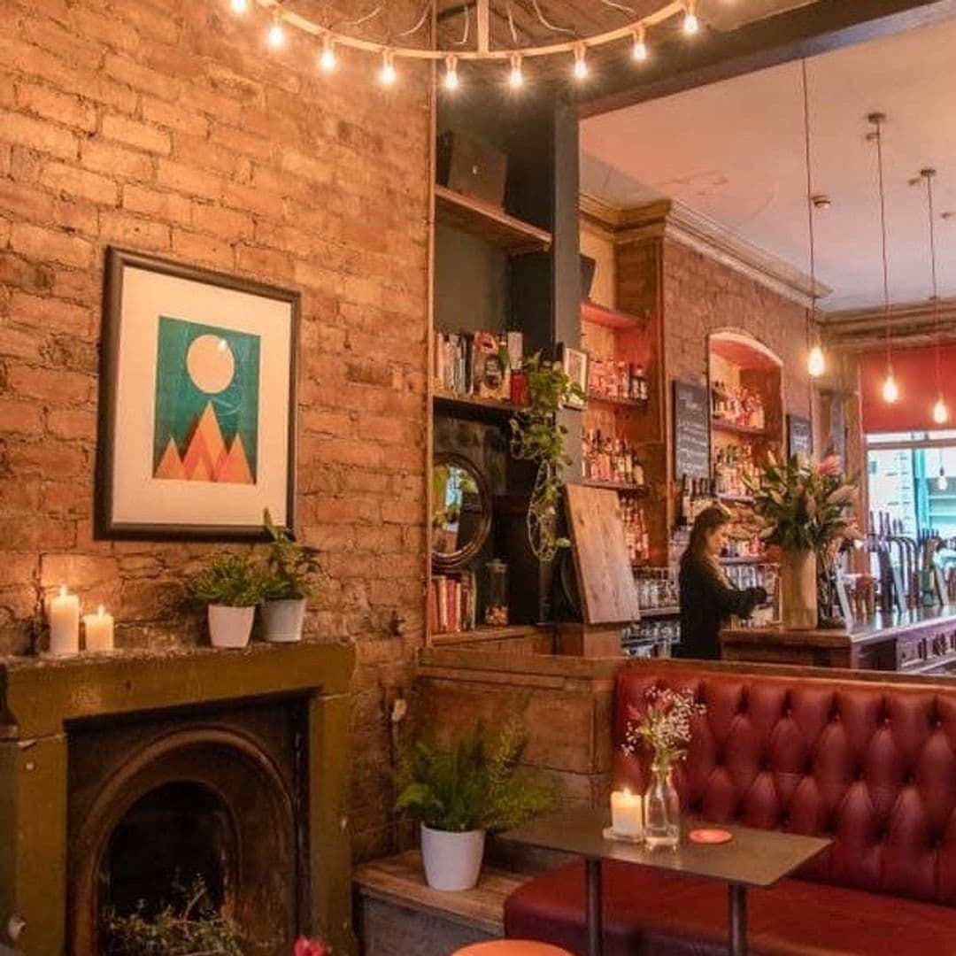 Rolands_Cozy_Cafe_Interior_Brick_Walls_String_Lights Interior of Roland's featuring exposed brick walls adorned with artwork, string lights, a cozy red booth, and a bar area with shelves of bottles, with a bartender in the background.