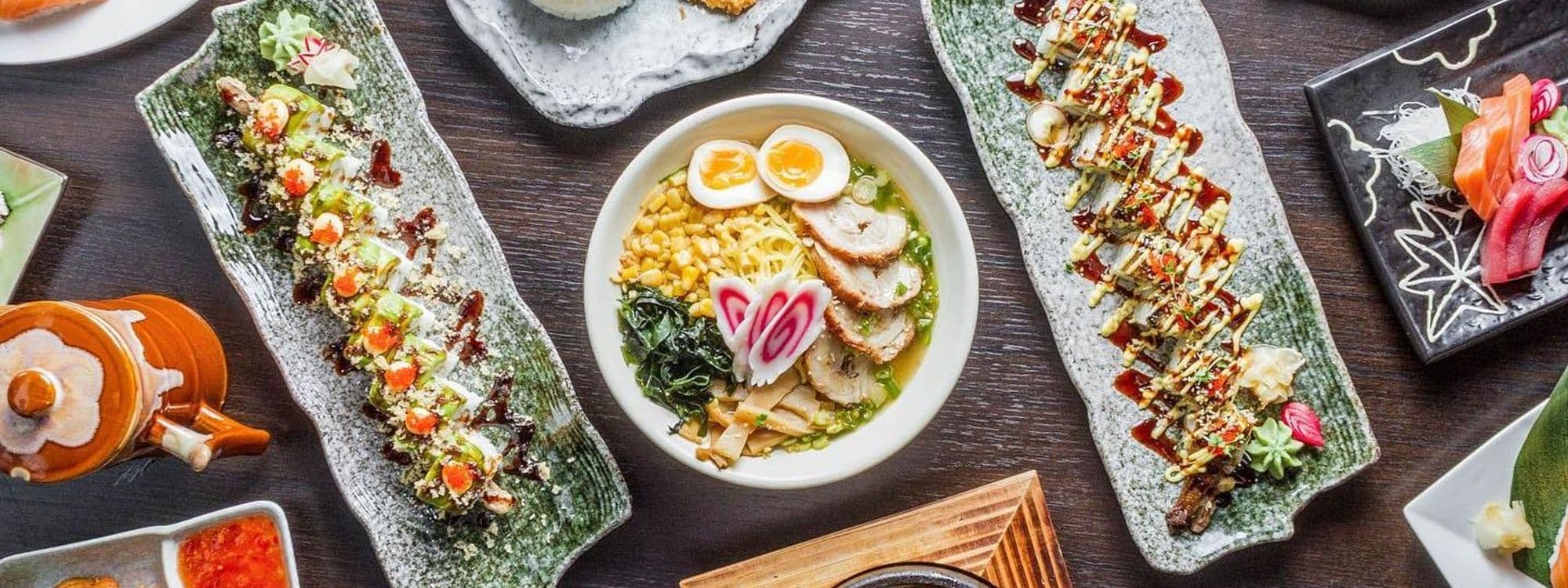 Assorted traditional Japanese dishes on a dark wooden table at Sushi Waka, featuring sushi rolls, ramen with egg, and sashimi.