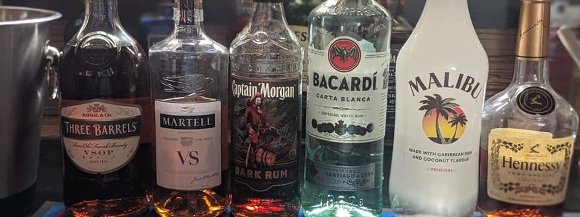 Assorted premium liquor bottles on display at The Grand Valley Bar & Kitchen, featuring brands like Three Barrels, Martell, Captain Morgan, Bacardi, Malibu, and Hennessy.
