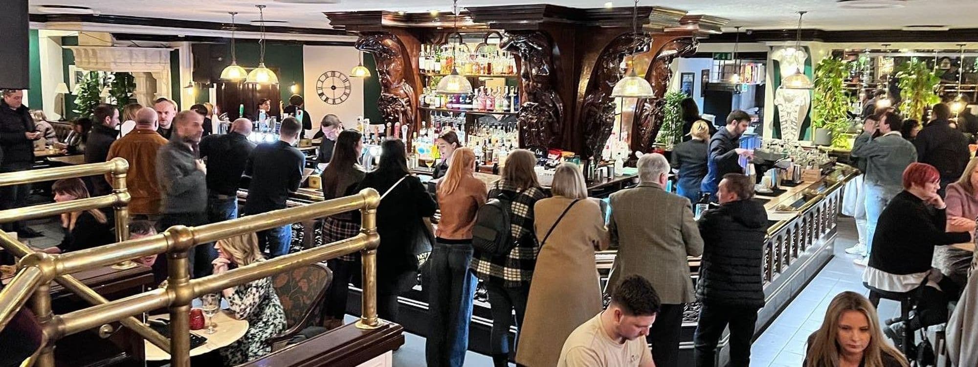 Interior of The New Conservatory showing patrons socializing and enjoying drinks at the bar with ornate wooden decor and plants in the background.