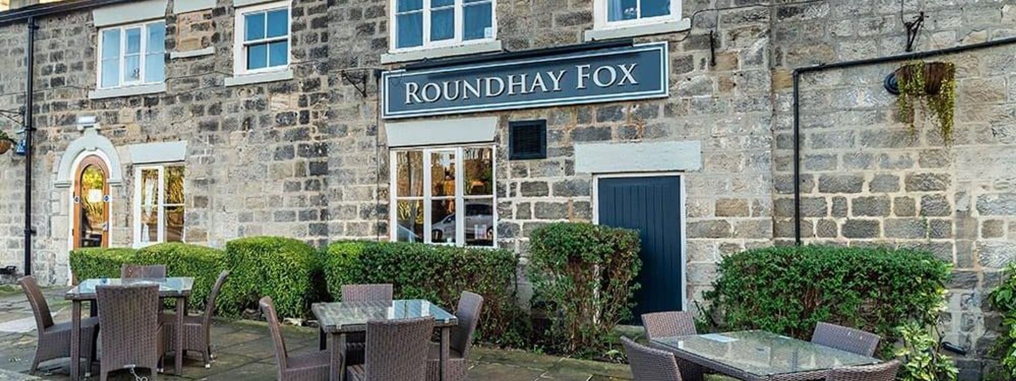 Exterior view of The Roundhay Fox, a traditional stone-built pub with a signboard, featuring outdoor seating and green hedges.