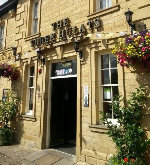 The Three Hulats Pub Entrance Floral Display Sunny Day Exterior view of The Three Hulats pub with a sandstone facade, adorned with colorful hanging flower baskets under a clear sky.