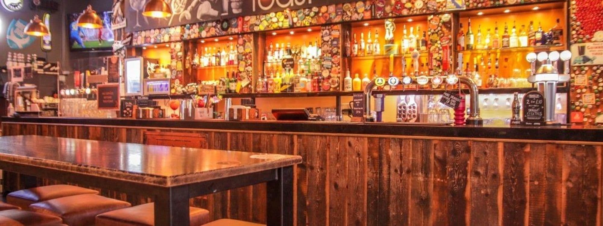 Interior view of Toast Bar featuring a well-stocked bar with a variety of bottles and taps, wooden bar counter, and seating area with leather stools and tables.